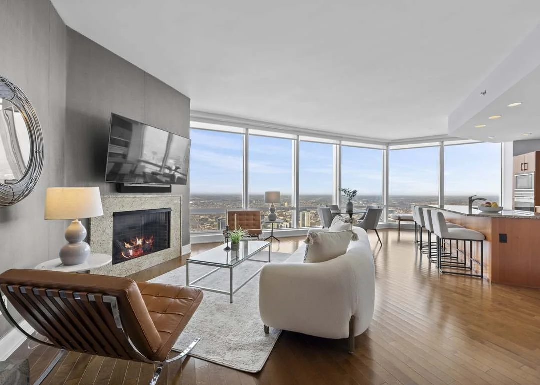 Modern living room with large windows offering a city view, fireplace, flat-screen TV, white sofa, armchair, glass coffee table, and bar stools at the kitchen island.