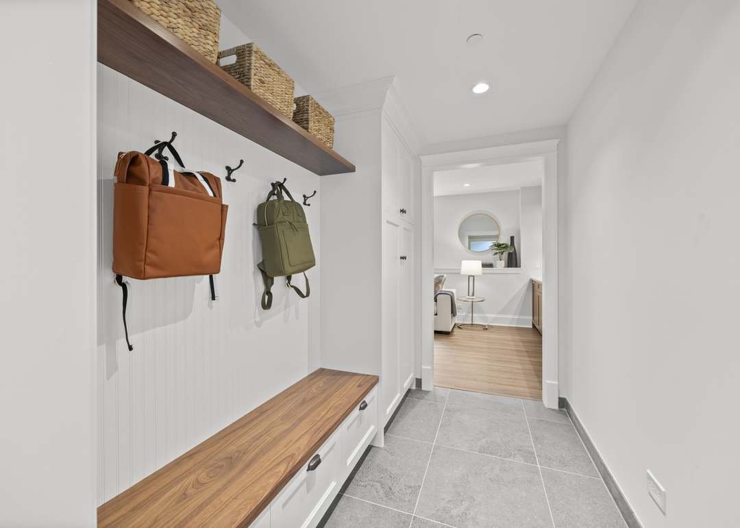 Entryway with white walls, a wooden bench with drawers, coat hooks with bags, and a wooden shelf with baskets, leading to a living room with a mirror, a lamp, and hardwood flooring.