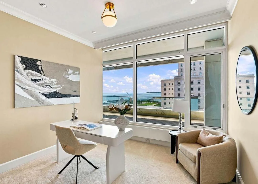 A modern living room with a large window view of a cityscape and water, beige walls and carpet, white desk with a chair, couch, lamp, and decorative vase.