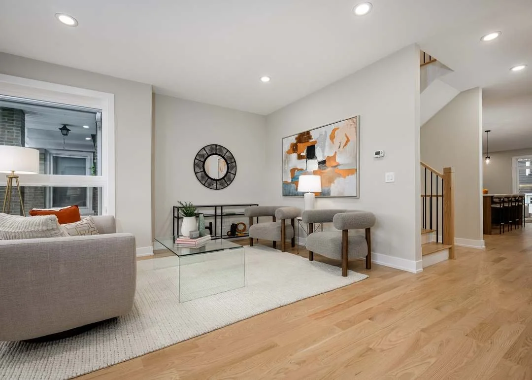 Modern living room with a light-colored sofa, a glass coffee table, a small black metal bench, abstract wall art, and a mix of neutral and wooden accents.