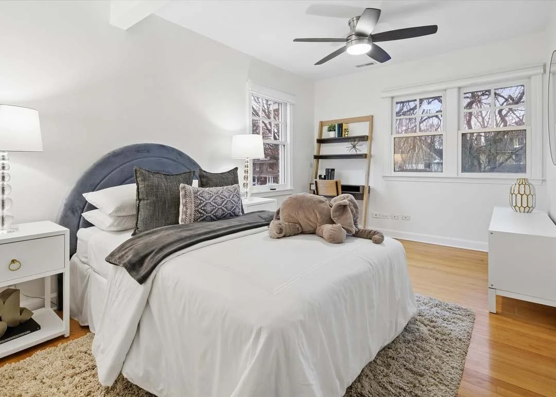 A bedroom with white walls, a bed with a gray upholstered headboard, white bedding, and a plush dog stuffed animal on top. There are nightstands with lamps on either side of the bed, a wooden bookshelf with a few books and decor items, and large windows showing trees outside. The room has hardwood floors, a beige rug, a ceiling fan, and a white dresser with a decorative vase.