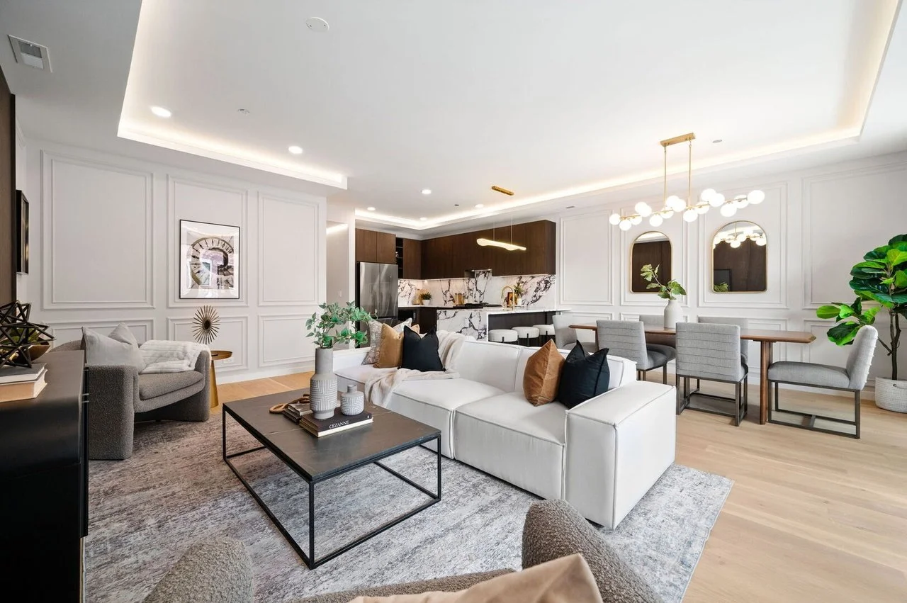 Modern open-concept living room and kitchen with white walls, wooden flooring, gray and white furniture, a black coffee table with decorative vases, a dining table with gray chairs, and green plants.