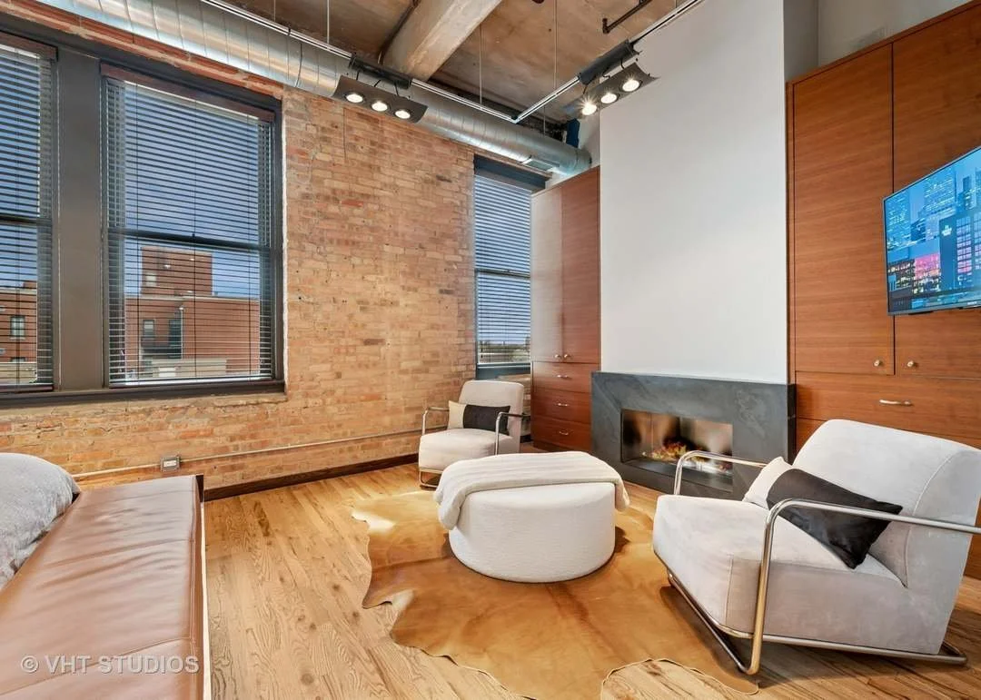 Modern living room with exposed brick wall, large windows with blinds, a fireplace, and minimalist furniture including a white armchair, a beige sofa, and a cowhide rug, with a mounted television on the wooden cabinets and an industrial ceiling with visible ductwork.