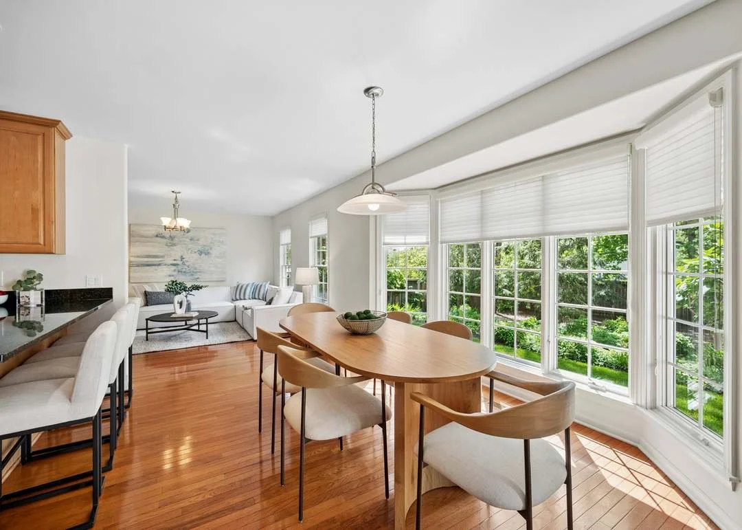 Bright dining area with large bay windows, wooden dining table with six chairs, and a view of green trees outside.