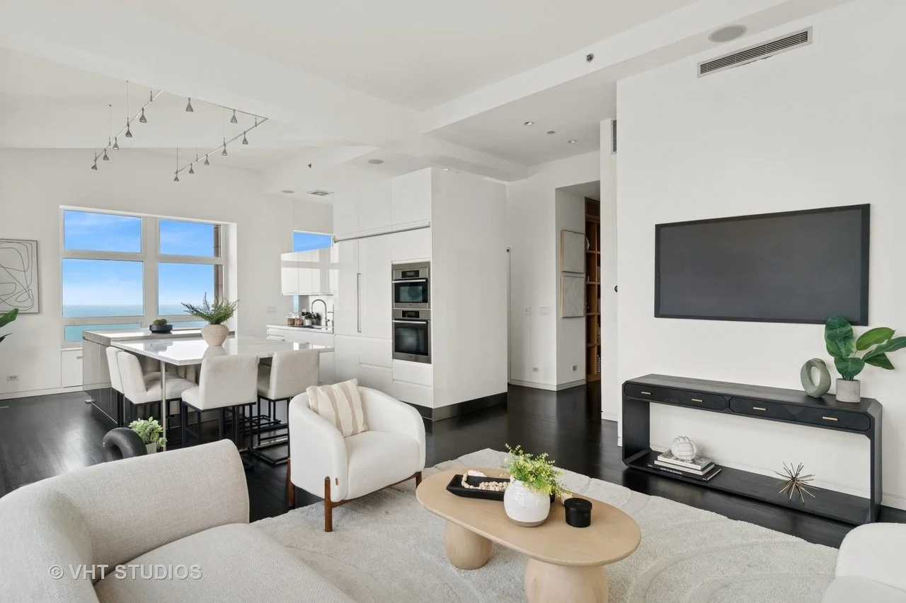 Modern living room and kitchen with white walls, black flooring, a wall-mounted TV, and large windows showing a view of the ocean.