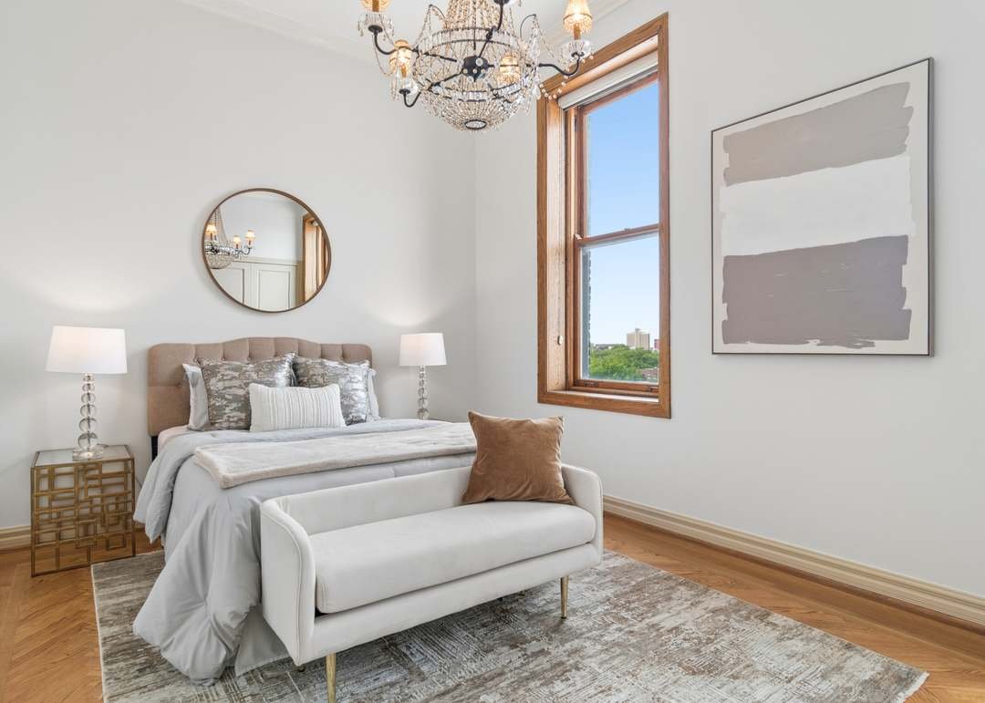 A bedroom with white walls, a beige upholstered bed with decorative pillows, two white nightstands with white lamps, a circular mirror above the bed, a window with natural wood trim showing a view of trees and buildings, a white sofa at the foot of the bed, a large abstract painting on the wall, a chandelier hanging from the ceiling, and a patterned area rug on the wooden floor.
