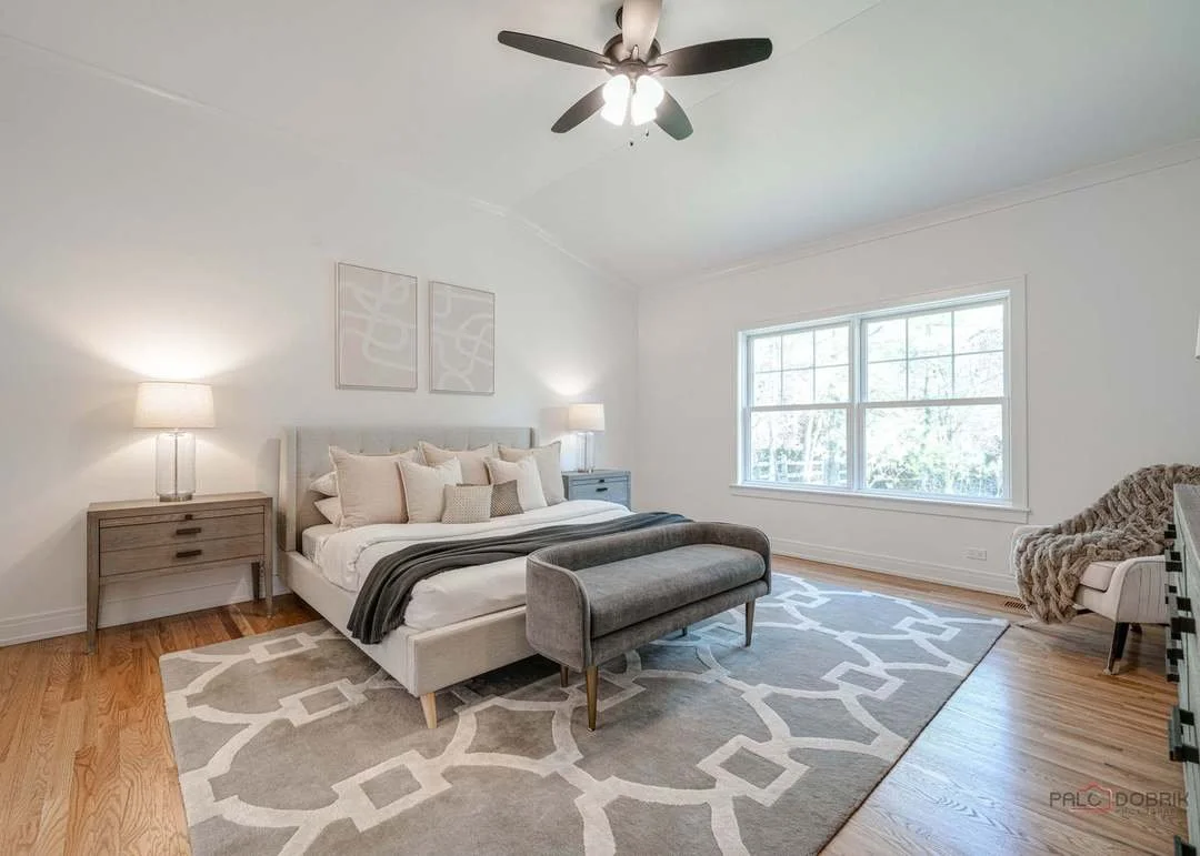 Modern bedroom with a large bed, gray upholstered bench at foot, gray nightstands with lamps, large window, white walls, hardwood floor, and a ceiling fan.