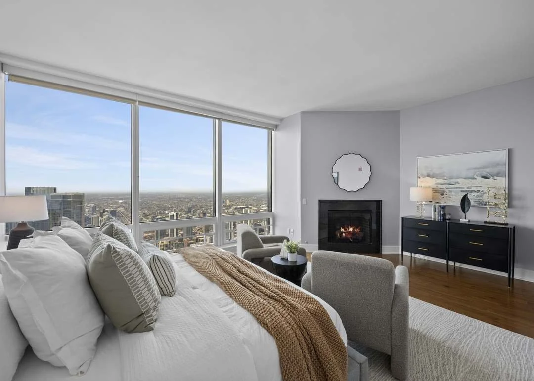 Living room with large windows offering city views, a bed with pillows and a blanket, a fireplace, a black sideboard with decorative items, a painting, and a chair.