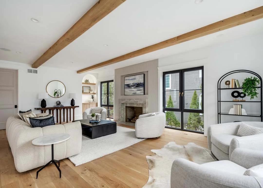 Living room with white sofas, a black coffee table, fireplace with a painting above, large windows, and a black metal shelf with decor, wood ceiling beams, and hardwood floors.