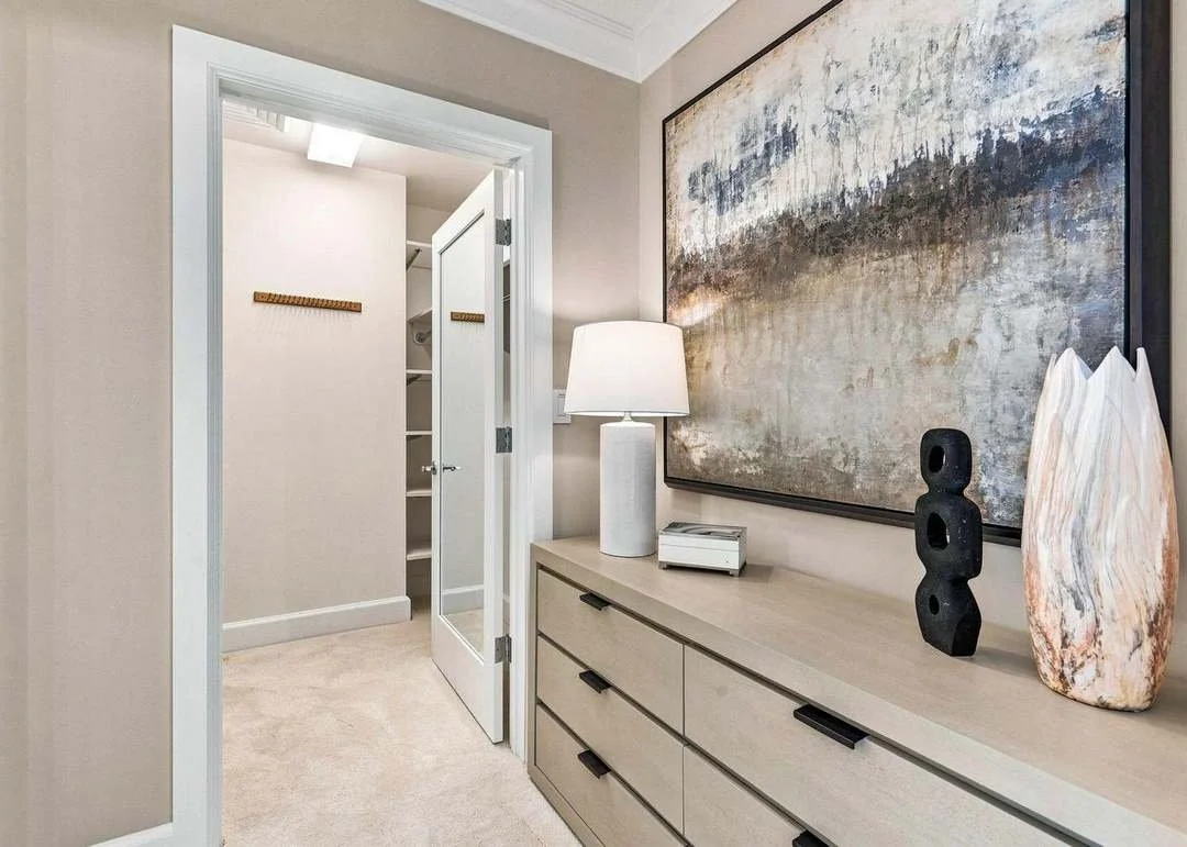 Interior view of a hallway with a dresser, table lamp, abstract art, decorative objects, and a closet in the background.