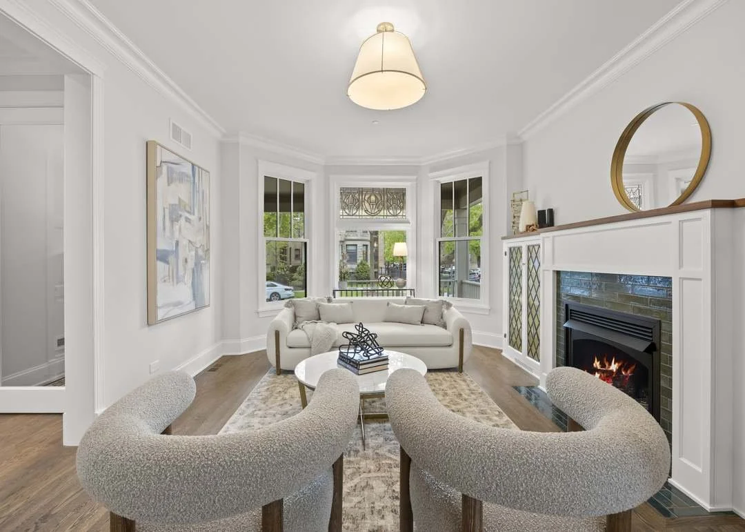Bright living room with a white sofa, two curved armchairs, a coffee table, a fireplace, and large bay windows, decorated in neutral tones.