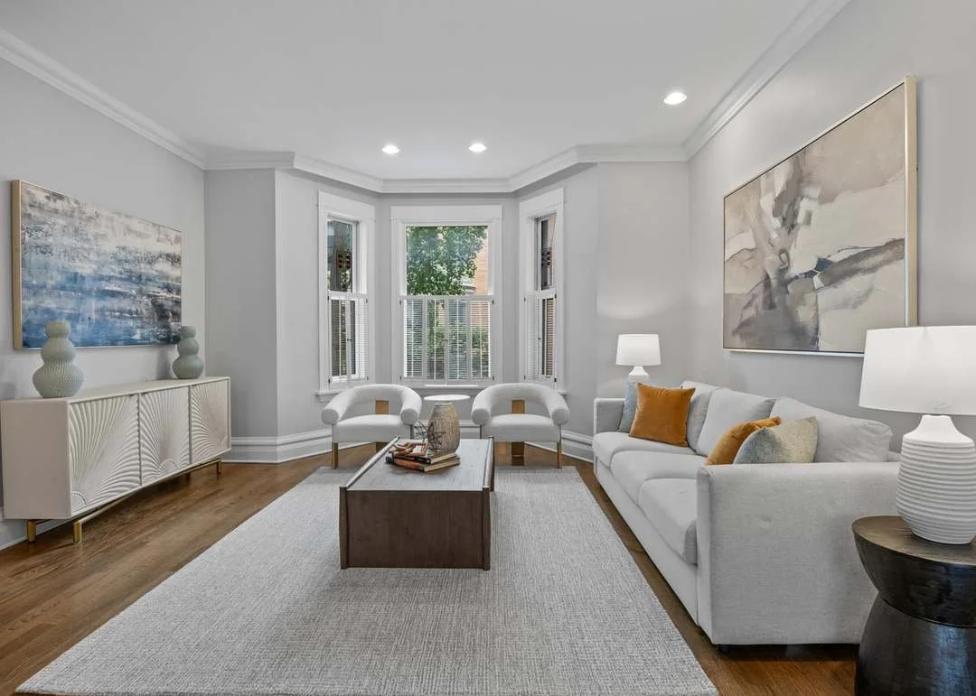 Bright living room with white walls, hardwood floors, a white sofa with pillows, two white armchairs near a bay window, a wooden coffee table, and abstract artwork on the walls, illuminated by ceiling and table lamps.