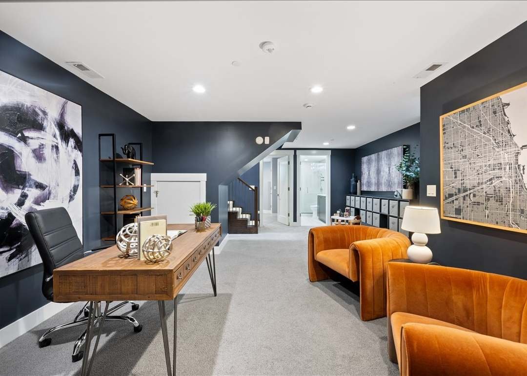 Modern home office with a black and white abstract painting, a wooden desk, black chair, orange armchairs, a white table lamp, wall art, and storage cabinets on navy-blue walls.