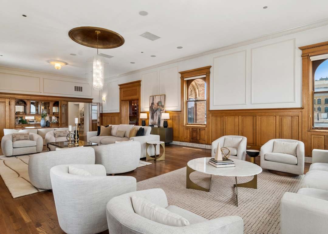 Spacious living room with white upholstered furniture, wooden wall paneling, large windows, and a modern coffee table.