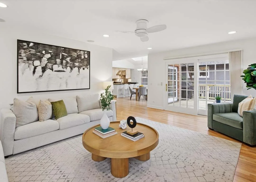 Bright living room with white sofa, green armchair, round wooden coffee table, and large window with sliding glass doors leading to a patio.