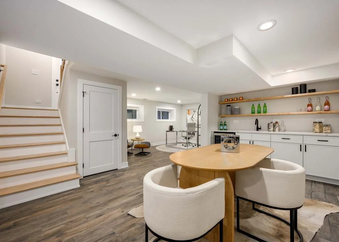 Modern basement with wooden flooring, white walls, open staircase, small seating area, and kitchen area with open shelves, white cabinets, and a wooden table with two chairs.
