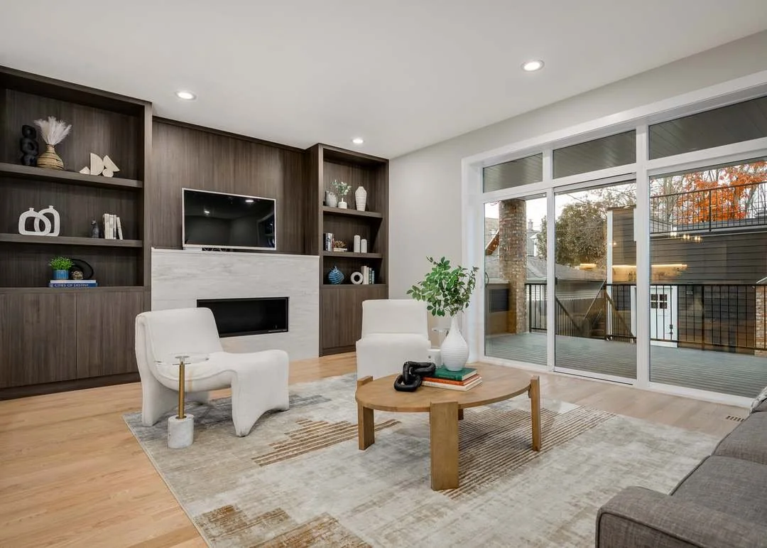 Modern living room with built-in wooden shelves, a fireplace, a flat-screen TV, white chairs, a wooden coffee table with decorative items, a gray sofa, and large glass sliding doors leading to an outdoor patio.