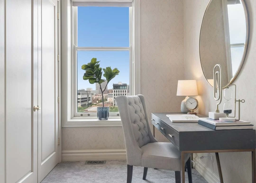 Home office with a desk, a tufted chair, a large round mirror, a table lamp, a clock, and a plant on the window sill, with buildings outside window.
