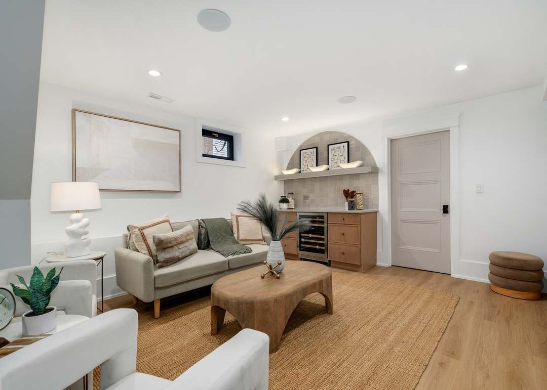 Living room with beige sofa, wooden coffee table, potted plant, wall art, table lamp, and built-in shelves with decorative items, featuring hardwood flooring and white walls.