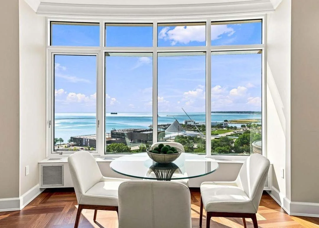 A dining area with a glass table and four white chairs, a bowl of green fruit on the table, and a large window showcasing a beach and ocean view with blue sky and clouds.