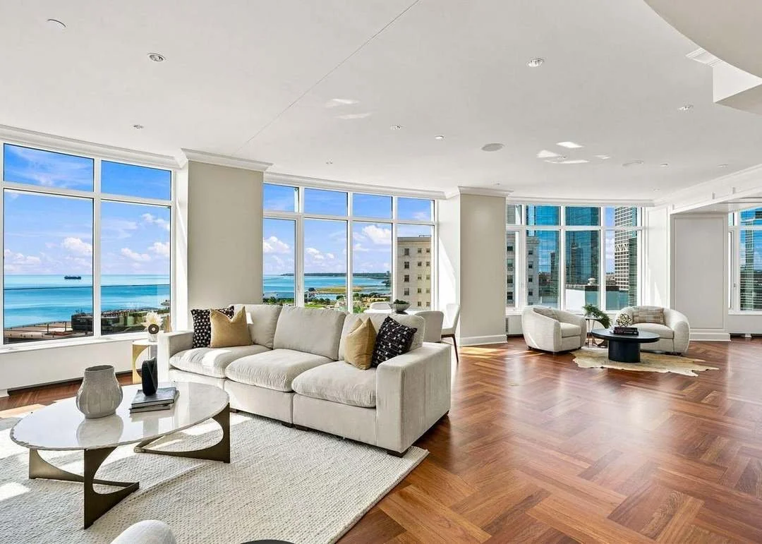 Bright, spacious living room with large windows offering ocean views, furnished with a white sofa, coffee table, armchair, and decorative pillows, with wood flooring and minimal decor.