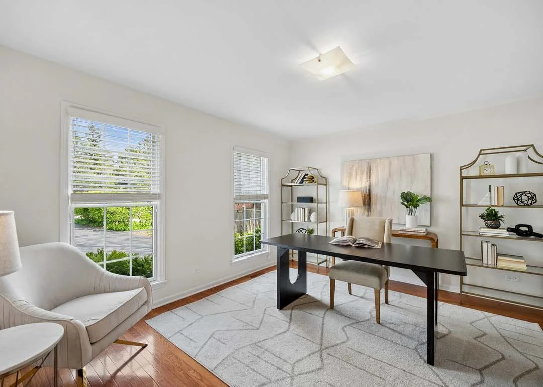 Bright, minimalist home office or living room with two large windows, a white couch, a dark wooden table, an upholstered chair, a beige armchair, a lamp, and metal shelving with decor and books.