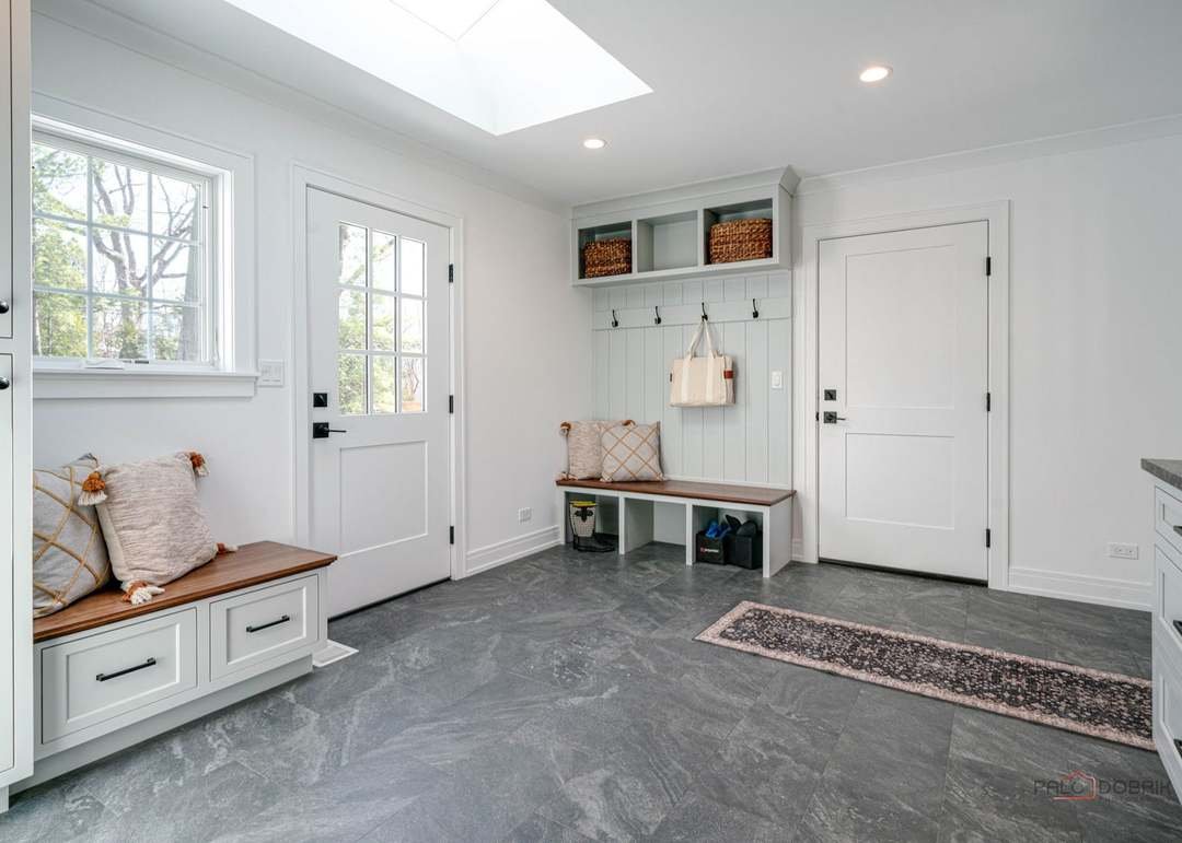 Entryway with white walls, a bench with pillows, a built-in storage with hooks, baskets, and a small rug on a gray tile floor.