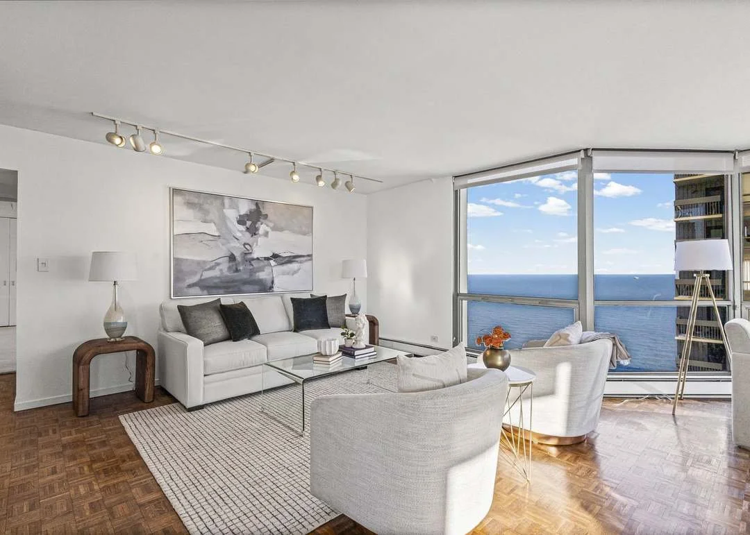 Living room with white sofa, white armchairs, glass coffee table, ocean view through large windows, wooden floor, modern lamps, wall art, and decorative pillows.