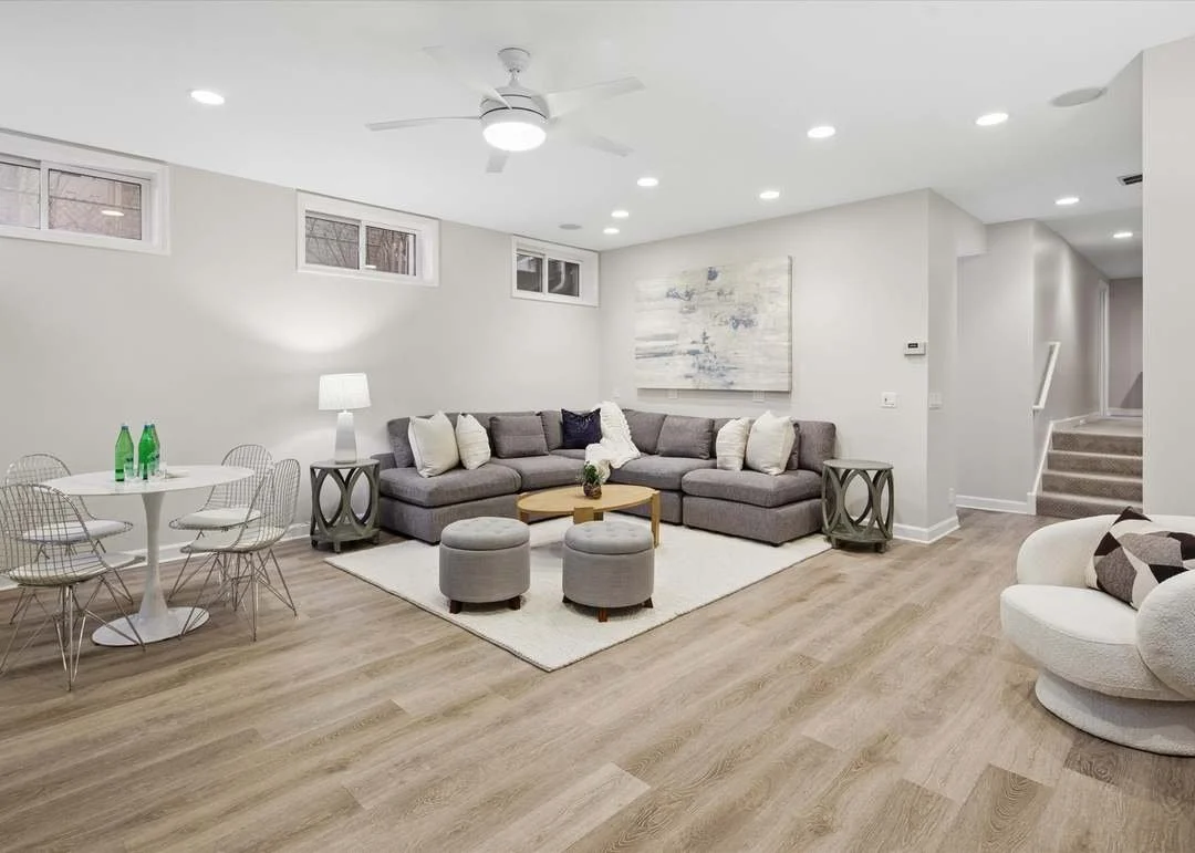 A modern living room with a large L-shaped gray sofa, a white rug, two gray ottomans, a wooden coffee table, and a white round lounge chair. There are multiple small windows, a ceiling fan, and recessed lighting. Decor includes a large abstract painting and cushions.