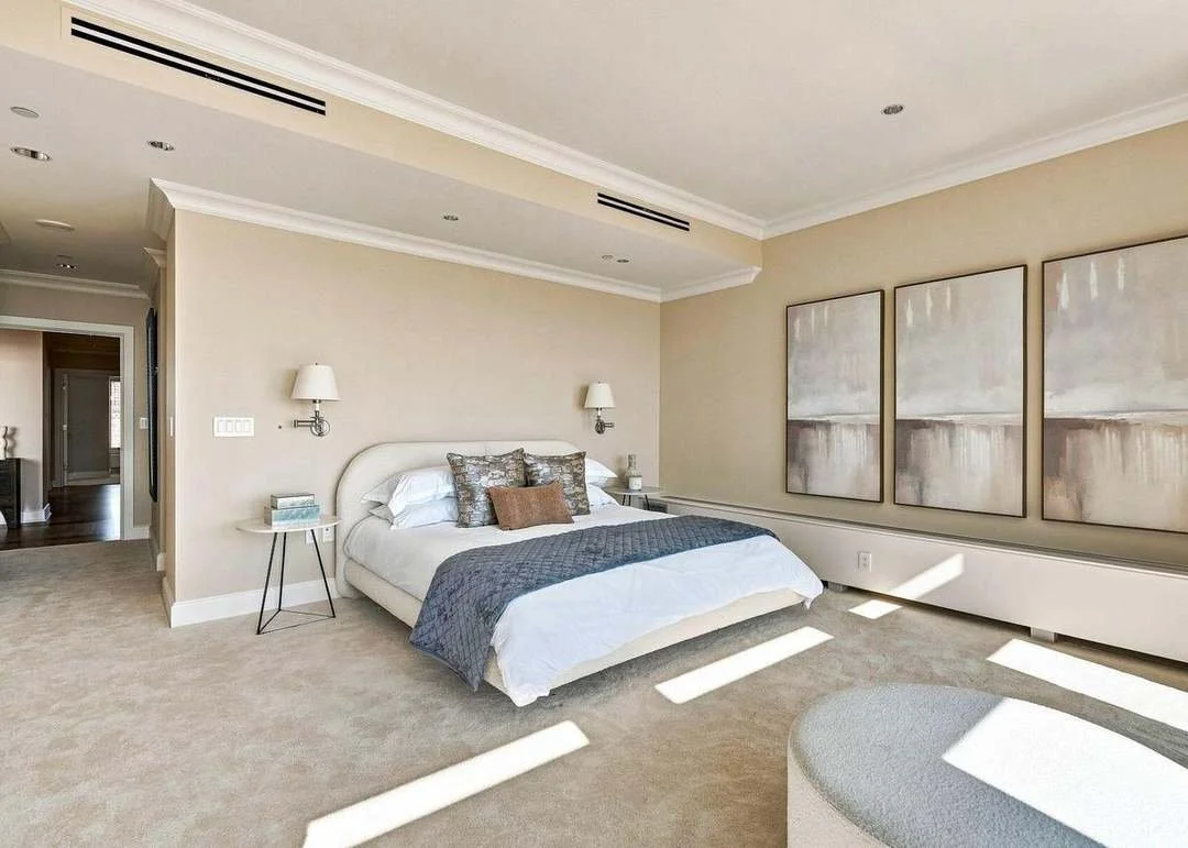 A modern bedroom with a white bed, blue and brown decorative pillows, gray quilt, side tables with lamps, and three large abstract paintings on the wall, illuminated by natural light.