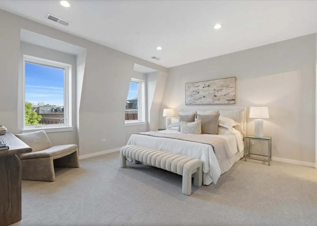 A bright, modern bedroom with white walls, a large bed with white bedding and several pillows, two nightstands with lamps, a bench at the foot of the bed, and two windows showing a cityscape with a blue sky.