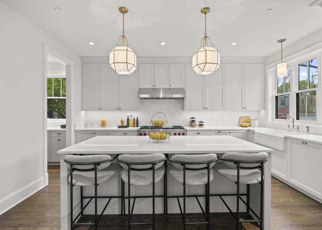 A modern white kitchen with a large island, four bar stools, and pendant lighting fixtures.