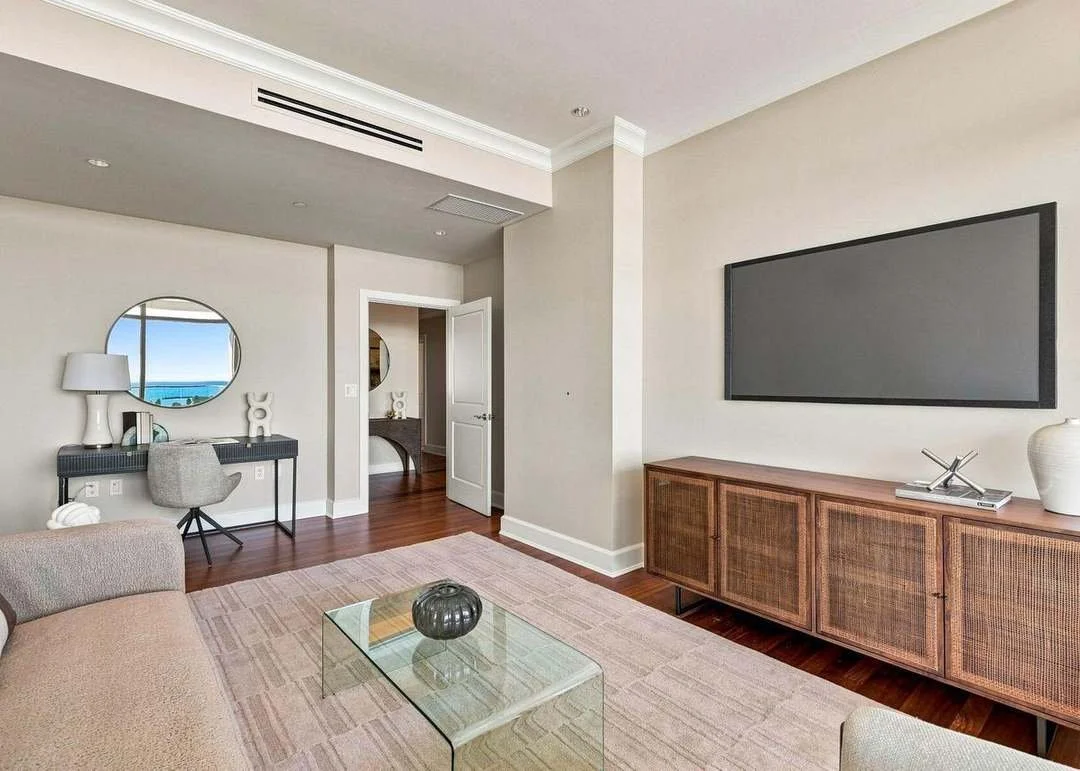 Living room with a beige sofa, glass coffee table with a dark decorative item, wooden sideboard with a flat-screen TV, and a small desk with a mirror and decorative objects.