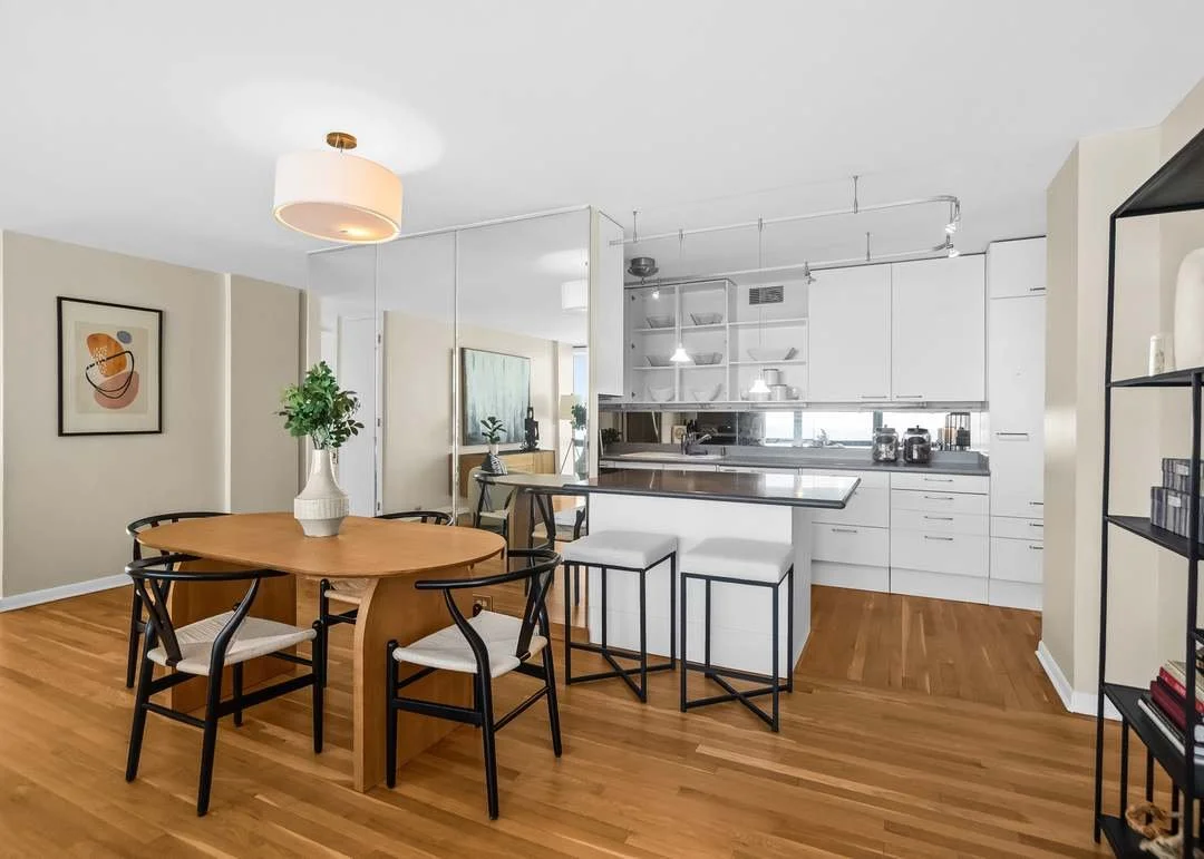 Open-concept dining area and kitchen with hardwood floors, a wooden dining table with five chairs, a white kitchen island with black metal stools, and white cabinets with open shelving.