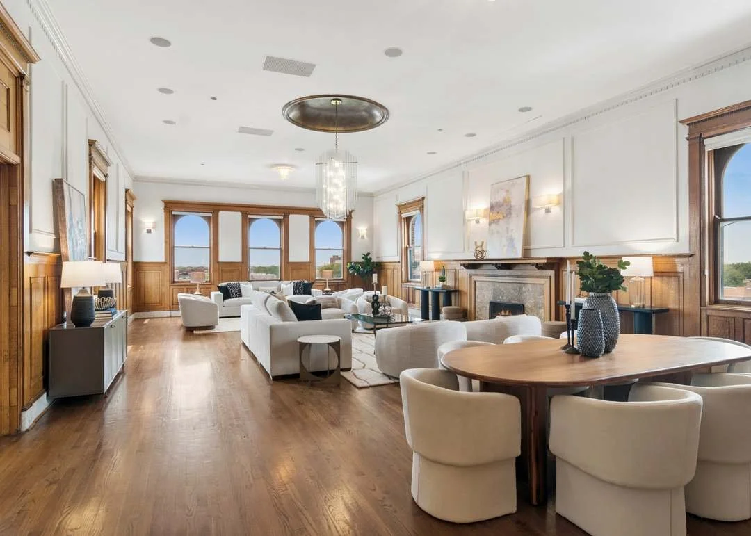 Spacious living room with white sofas, a wooden dining table with beige chairs, wooden wall paneling, large windows, a fireplace, and elegant decor.