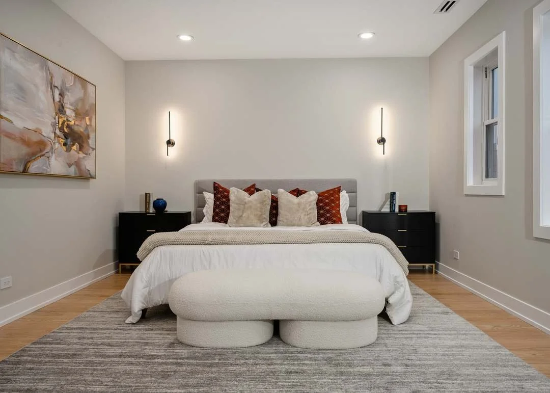 A modern bedroom with a white bed, multiple pillows, black nightstands, wall-mounted lighting, a gray upholstered headboard, and abstract artwork on the wall. The room has hardwood floors, a large window, and neutrally colored decor.