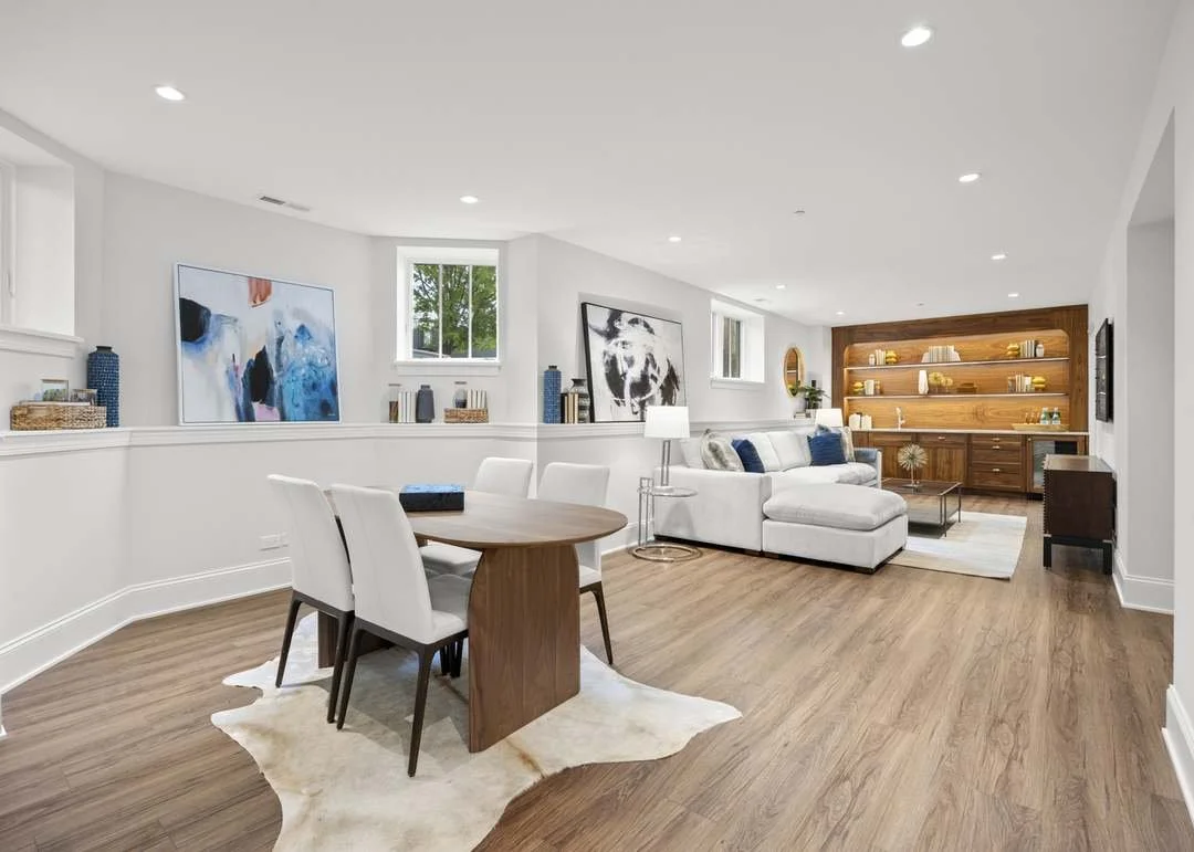 Bright living room with white walls, wooden floor, white sectional sofa with blue pillows, wall-mounted TVs, built-in wooden shelves with decor and books, oval dining table with white chairs, and large artwork on the walls.