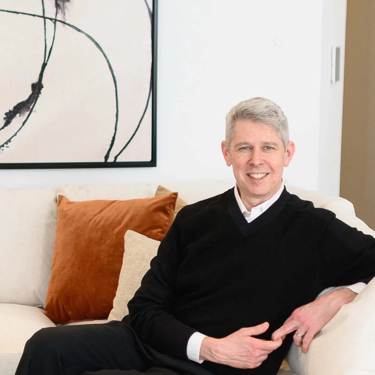 A middle-aged man with gray hair sitting on a cream-colored sofa with brown and beige pillows, smiling at the camera in a modern living room, with abstract wall art in the background.