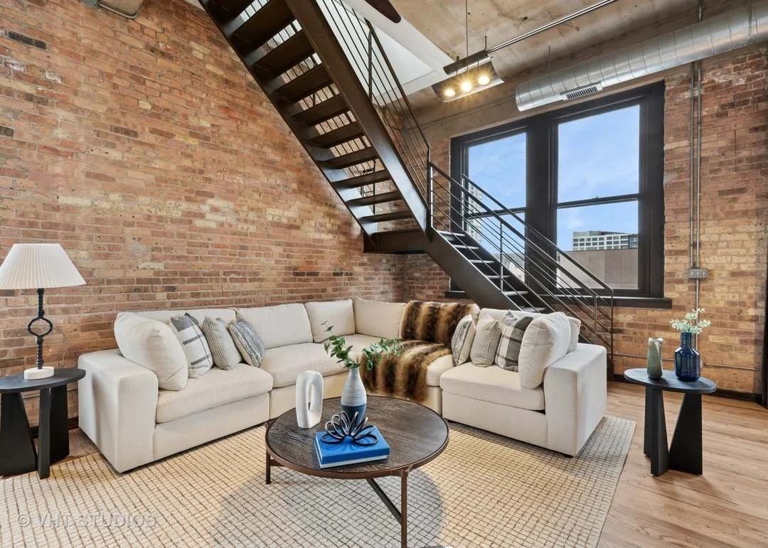Living room with a white sofa, black coffee table, brick wall, large window, staircase, and industrial ceiling in a modern apartment.