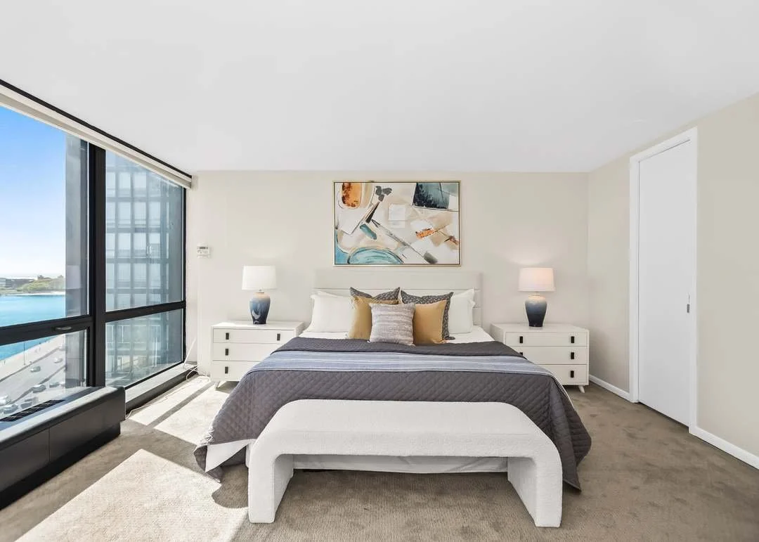 Modern bedroom with a large window overlooking water, white walls, a bed with gray and tan pillows, and two nightstands with lamps.