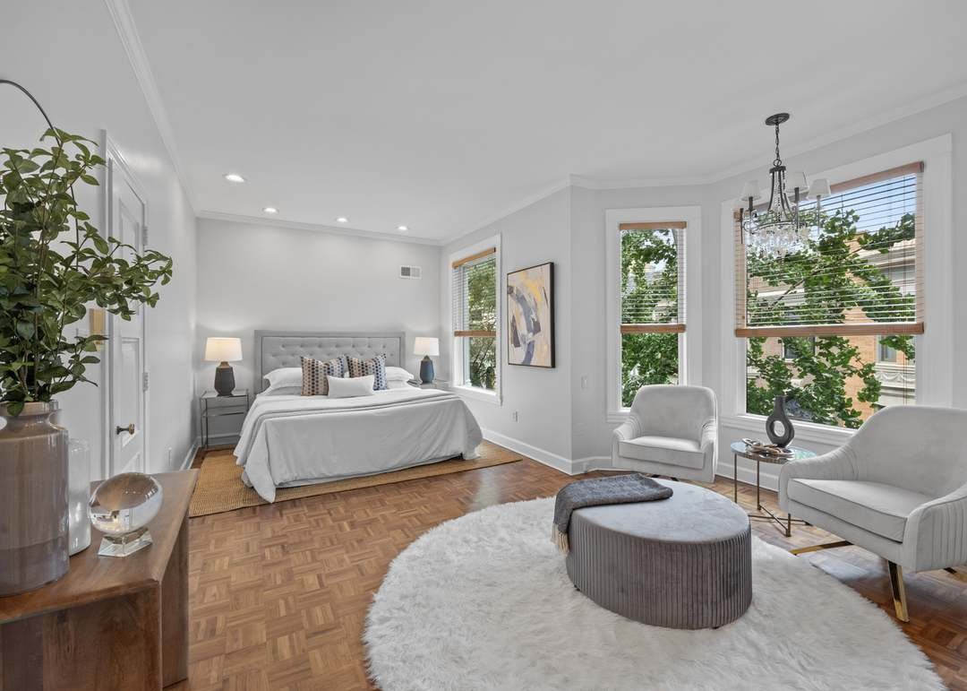 Bright bedroom with white walls, hardwood floor, bed with gray headboard, white bedding, and decorative pillows. Two nightstands with lamps, a large window, and white chairs with a small table near the windows. A chandelier hanging from the ceiling, a white shag rug on the floor, and a wooden dresser with decorative items.