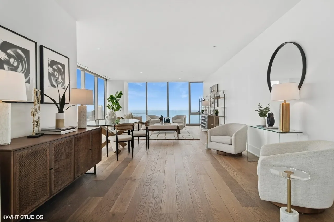 Modern living room with large floor-to-ceiling windows offering a view of the ocean, featuring white walls, wooden flooring, and contemporary furniture including armchairs, a sofa, a coffee table, and decorative items.