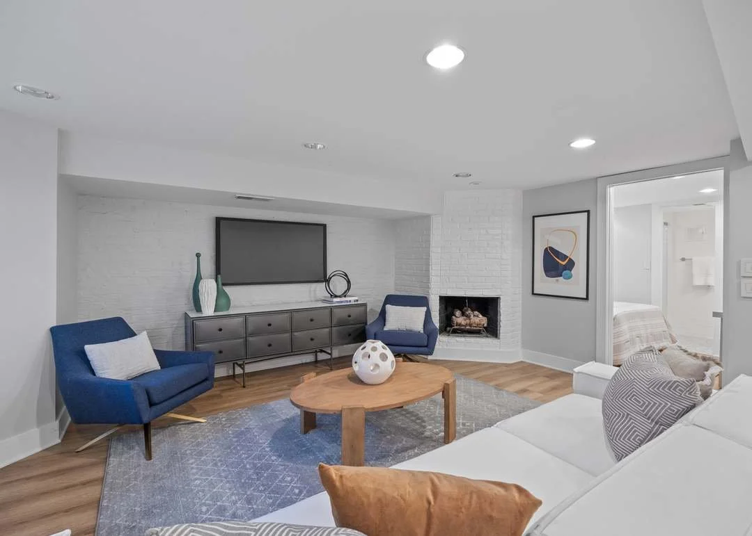 Living room with white walls, hardwood floors, a white brick fireplace, a white sofa with patterned pillows, two blue armchairs, a wooden coffee table with a decorative sphere, a gray media console, and framed artwork on the wall.