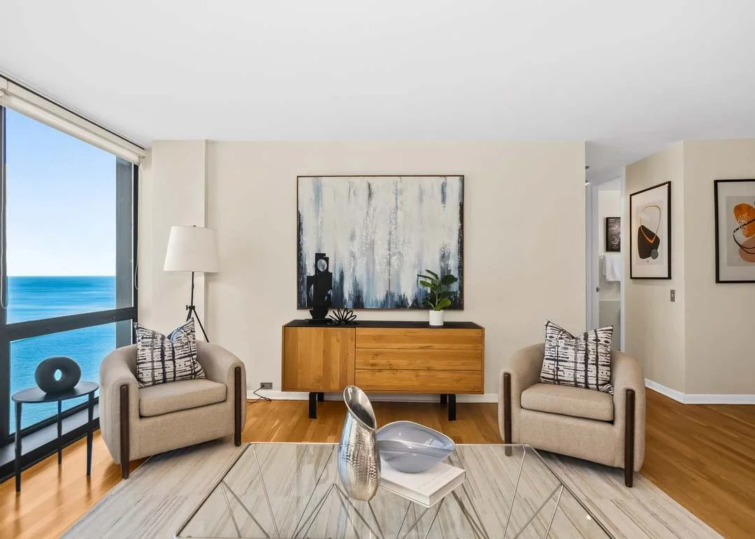 Modern living room with beige armchairs, a glass coffee table, abstract artwork on the walls, a wooden sideboard, and a large window showing an ocean view.