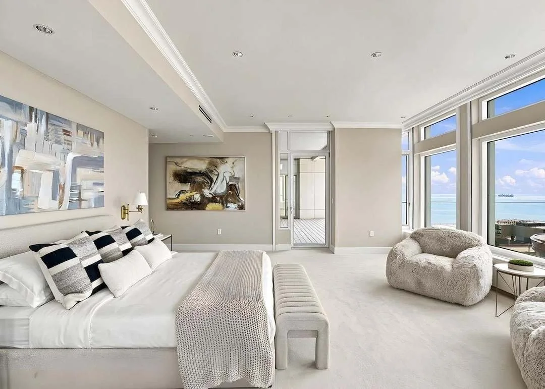 Modern bedroom with large windows overlooking the ocean, beige walls, abstract artwork, and contemporary furniture.
