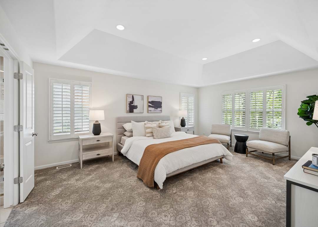 A spacious, brightly lit bedroom with a large bed, two nightstands with lamps, and seating area with chairs, in a neutral color scheme.