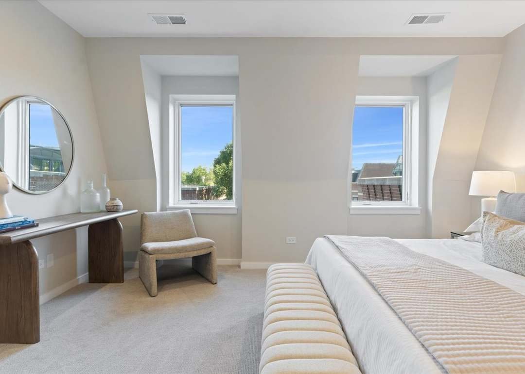 Light-colored bedroom with two windows, a bed with beige bedding, a bedside lamp, a beige chair, a wooden table with decorative objects, and a round mirror on the wall.