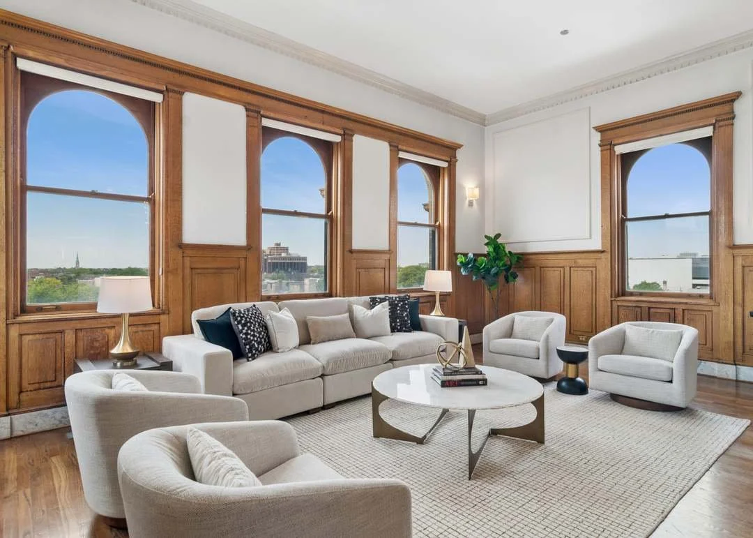 Living room with four large windows, wooden paneling, white walls, a white sofa with black and beige pillows, three beige armchairs, a round marble coffee table, a potted plant, and two lamps with white shades.