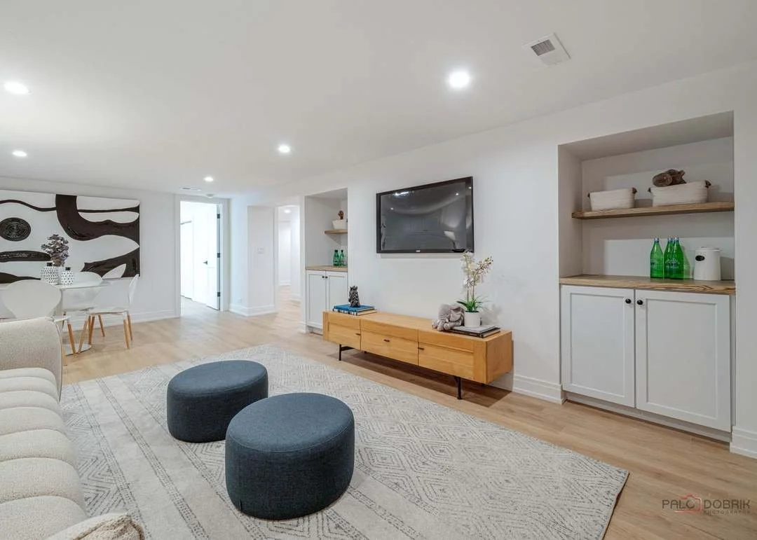Modern living room with a beige sofa, two dark blue ottomans, a wooden TV stand, and a flat-screen TV on a white wall. Open shelving with decorative items and green bottles, hardwood floor, and a dining area with white chairs and a round table in the background.