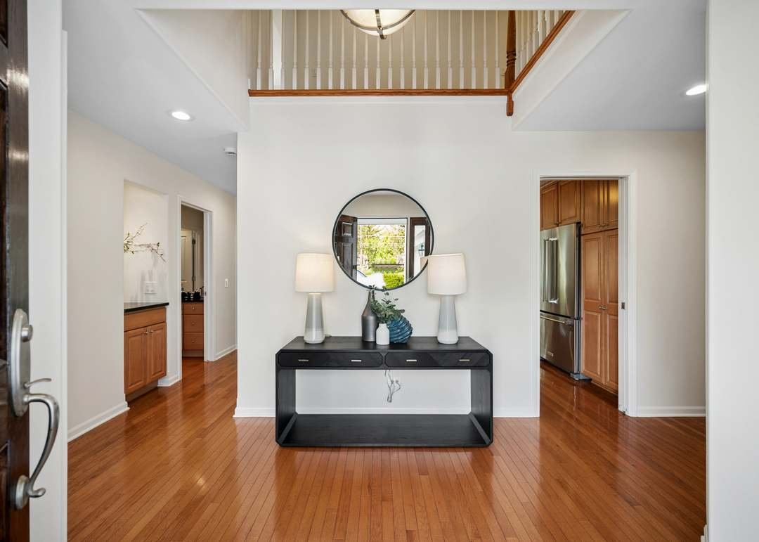 Interior view of a house entryway with hardwood floors, a black console table with two table lamps and decorative items, a round mirror above, and a glimpse of the kitchen with wooden cabinets to the right.